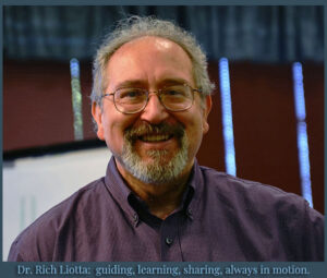 Dr. Rich Liotta smiling, wearing a purple shirt, standing in front of a presentation screen.